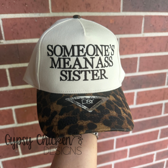 'SOMEONE'S MEAN ASS SISTER' embroidered natural and leopard hat held against a brick wall, brand 'Gypsy Chicken Designs' visible.