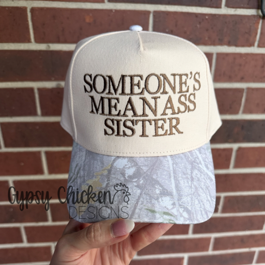 Cap with 'SOMEONE'S MEANASS SISTER' text held in front of a brick wall, brand 'Gypsy Chicken Designs' visible.